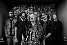 Layla Zoe Schwarz-Weiß Foto der fünfköpfigen Band Layla Zoe vor einer Wand mit zahllosen Stickern und Aufklebern