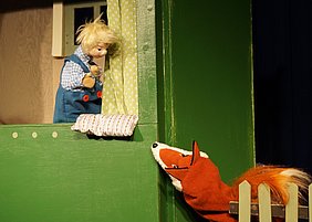 Ausschnitt aus dem Puppentheater Krümelchens Abenteuer, bei dem die Puppe am Fenster steht und zum orangen Fuchs hinaus sieht