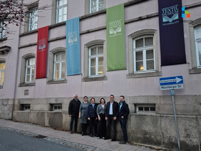 6 Personen, darunter 3 Männer und 3 Frauen, die das Projekt der neuen Fahnen am Textilmuseum verantworten stehen unter den bunten Fahnen an der Außenfassade des Helmbrechtser Textilmuseums.