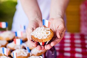 Frauenhände halten im Vordergrund einen Muffin, in dem eine französische Flagge steht. Im Hintergrund sind weitere Muffins zu erkennen.