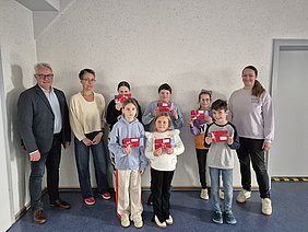 Gewinner des Luftballon-Wettbewerbs zum Wiesenfest Sechs Kinder stehen in der Mitte zwischen zwei ihrer Lehrinnen und dem Helmbrechtser Bürgermeister. In der Aula, vor einer weißen Wand. Sie halten ihre Gewinne in die Luft.