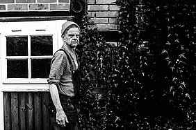 Der Singer-Songwriter abgelichtet vor einer Hausfassade mit Fenster und Begrünung im Hintergrund. Das Bild ist schwarz-weiß