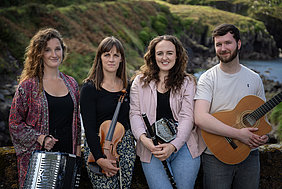 Drei Frauen und ein Mann stehen mit ihren Musikinstrumenten an einer Mauer. Sie blicken lächelnd in die Kamera. Im Hintergrund ist die Felsen- und Klippenlandschaft mit dem Anschluss zum Meer zu erahnen.