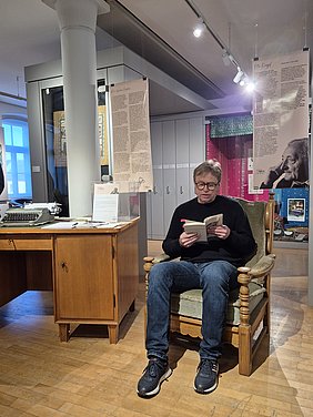 Reinhard Witzgall trägt die Geschichte "Das Kaffeekränzchen" vor Ein Mann sitzt auf dem original Sessel von Otto Knopf und liest zur Vernissage der Sonderausstellung seiner Ehren die Geschichte das Kaffeekränzchen