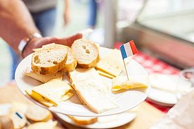 Auf einem Teller sind Baguettestücke und verschiedene Käsesorten angerichtet. In einem Käsestück steckt eine kleine französische Flagge.