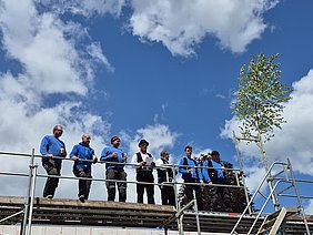 Andreas Tretter beim Richtspruch Auf dem Gerüst, neben dem Richtbaum, stehen unter blauem Himmel Andreas Tretter und seine Mitarbeiter der Zimmerei Tretter und halten Gläser in ihrer Hand. Andreas Tretter spricht den Richtspruch.