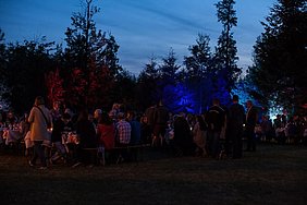 Abendliche Stimmung. Neben den in rot-weiß-blau angestrahlten Bäumen, sind viele Besucher zu sehen, die auf dem Areal stehen, auf den Biertischgarnituren sitzen und einen geselligen Abend verbringen.