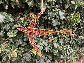 Ein aus Weidenästen geflochtener Vogel vor einer grünen Hecke