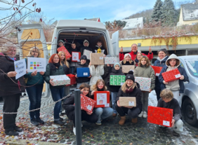Weihnachtspäckchen Aktion Eine große Gruppe an Schülerinnen und Schülern stehen mit Ihren Lehrkräften vor und zum Teil in einem Sprinterbus und halten eingepackte Weihnachtsgeschenke in die Luft.