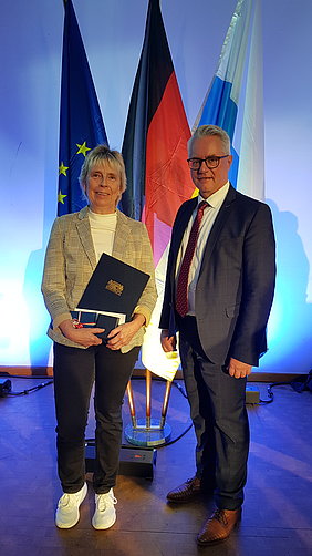 Uta Reif mit Bürgermeister Stefan Pöhlmann während der Feierstunde in Nürnberg Vor einem blauen Hintergrund stehen drei Fahnen, die europäische, die deutsche sowie die bayerische Fahne. Wiederum davor stehen eine Frau, die mit dem Ehrenamtspreis ausgezeichnet wurde, und der Helmbrechtser Bürgermeister.