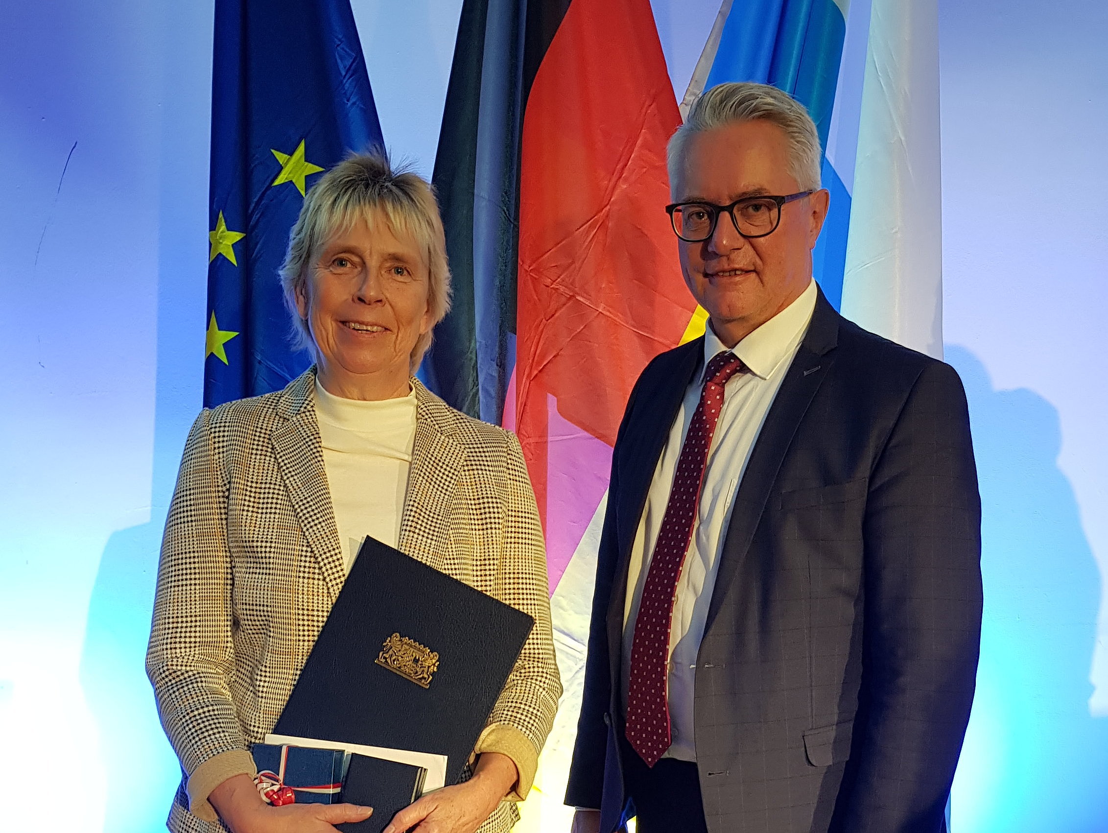 Vor einem blauen Hintergrund stehen drei Fahnen, die europäische, die deutsche sowie die bayerische Fahne. Wiederum davor stehen eine Frau, die mit dem Ehrenamtspreis ausgezeichnet wurde, und der Helmbrechtser Bürgermeister.