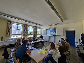 Architekt Martin Beyer stellt das Projekt vor 10 Personen sitzen in einem Klassenzimmer an in U-Form stehenden Tischen und folgen der Projektpräsentation von Architekt Martin Beyer.