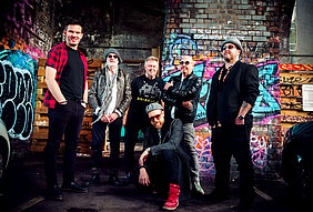 Die sechsköpfige Climax Blues Band auf einem Gruppenfoto unter einer Unterführung, deren gemauerten Wände mit Graffiti besprüht sind.