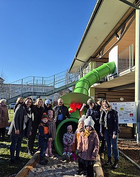 Die Projektverantwortlichen stehen bei strahlendem Sonnenschein an der neuen grünen Kinderrutsch der Kita Emilio. Ein Kind schneidet das Band durch.