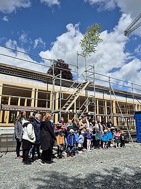 Eltern, Kinder und Erzieher singen das Handwerkerlied Einige Kinder singen gemeinsam mit ihren Eltern und Erziehern das Handwerkerlied. Sie stehen dabei im zukünftigen Außenbereich des Kindergartens. Über ihnen thront der Richtbaum vor blauem Himmel.