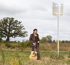 Orillia Der Sänger Orillia steht mit seiner Gitarre in der Natur, umgeben von Bäumen und Grasflächen