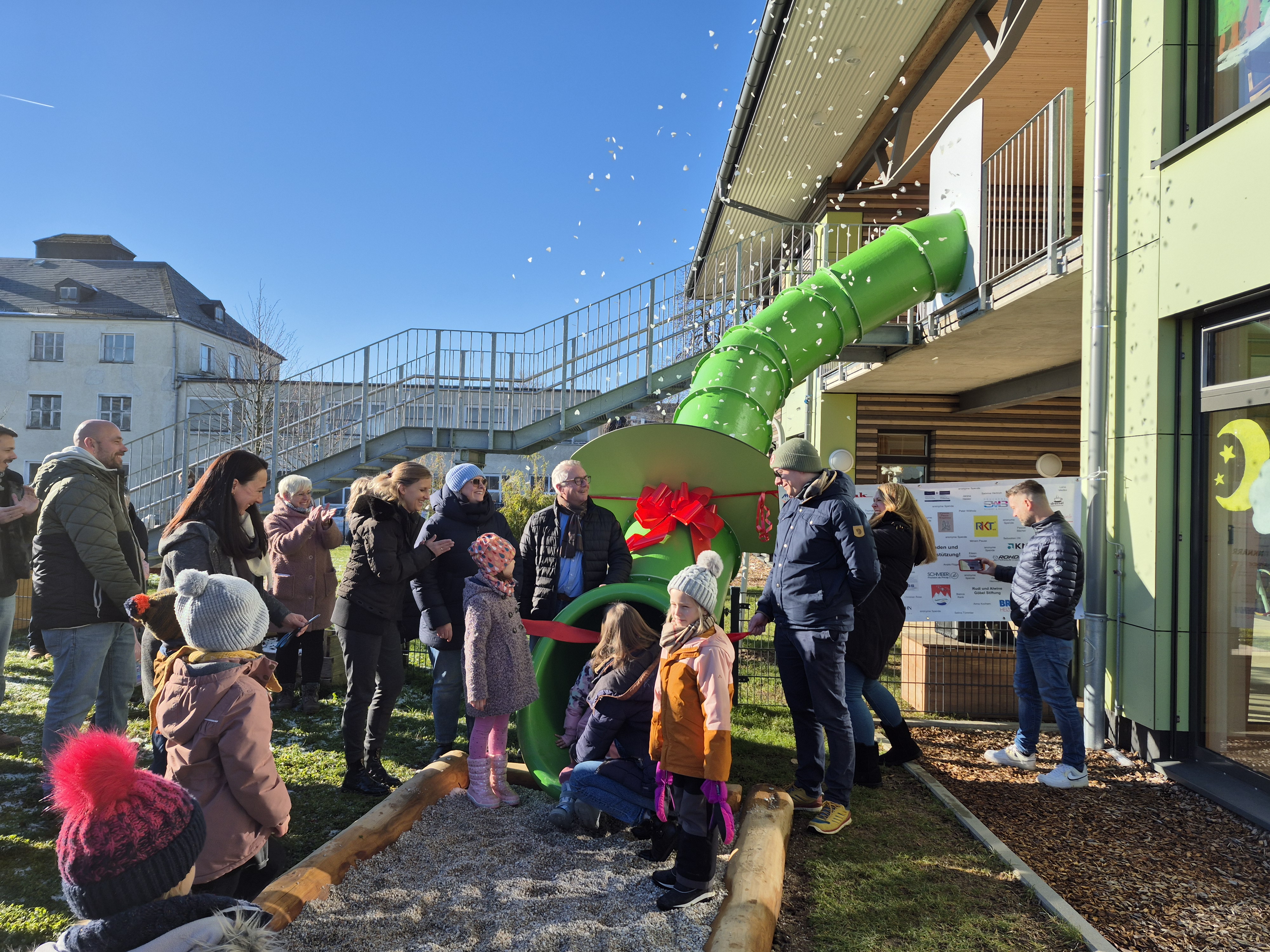 Die Projektverantwortlichen stehen bei strahlendem Sonnenschein an der neuen grünen Kinderrutsch der Kita Emilio. Ein Kind schneidet das Band durch.