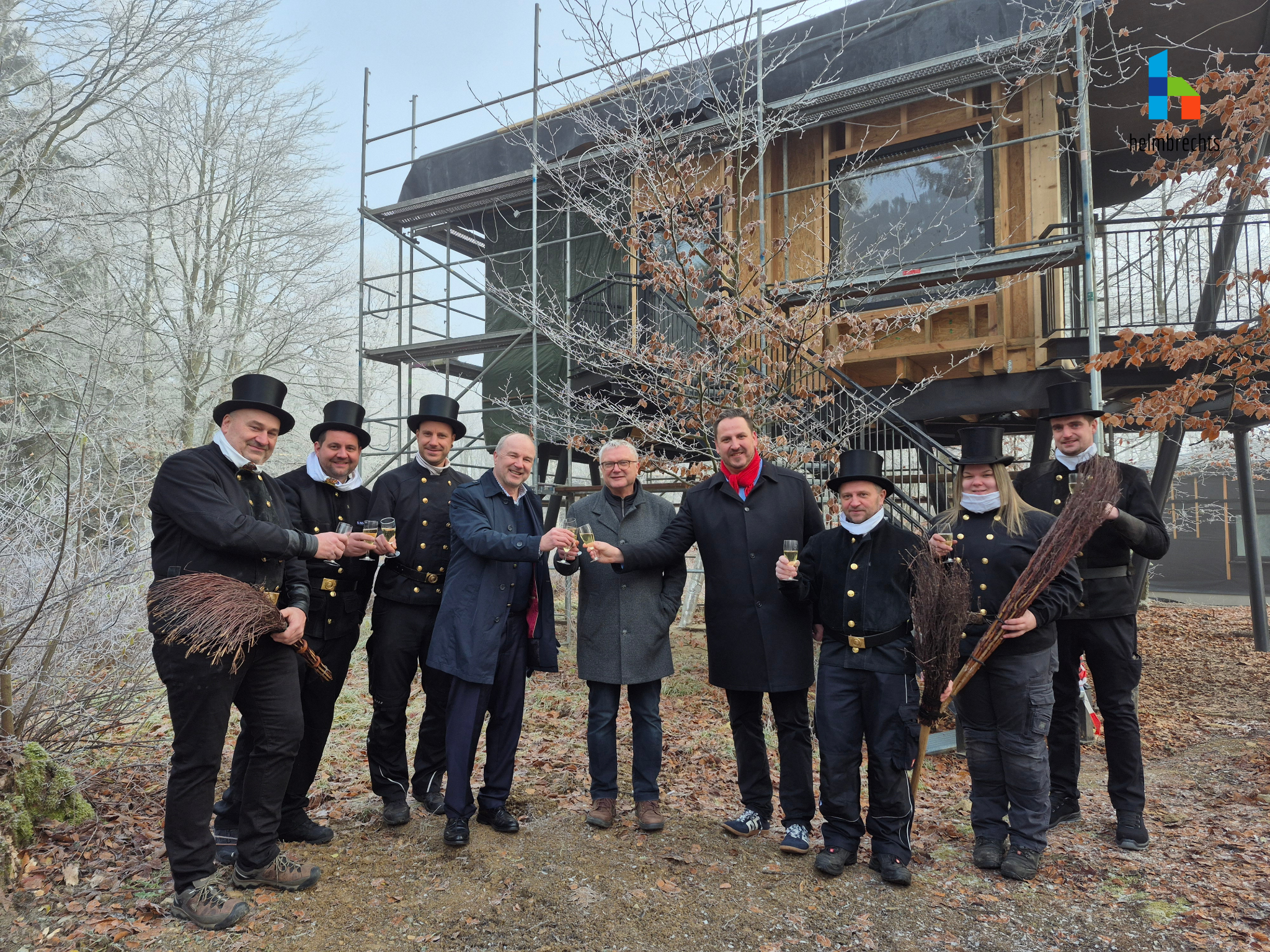 Neun Personen stehen mit Sektgläsern in der Hand vor einem Baumhaus am Kirchberg und stoßen bei winterlichem Sonnenschein auf das neue Jahr an. Darunter sind erster Bürgermeister der Stadt Münchberg, zweiter Bürgermeister der Stadt Helmbrechts und die Kaminkehrer aus beiden Städten