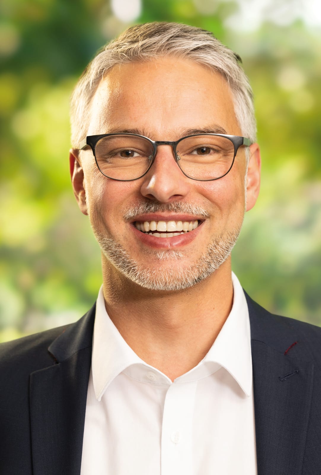 Bürgermeister Dr. Pascal Bächer Bürgermeister Pascal Bächer im schwarzen Anzug mit weißem Hemd vor einem grün bewachsenen, verschwommenen Hintergrund