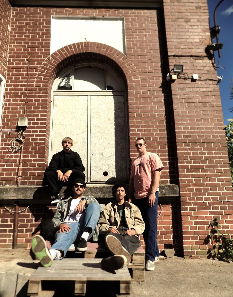 Vier Männer der Rolling Chocolate Band stehen vor einem Gebäude mit roten Backsteinen in der Sonne und blicken in die Kamera