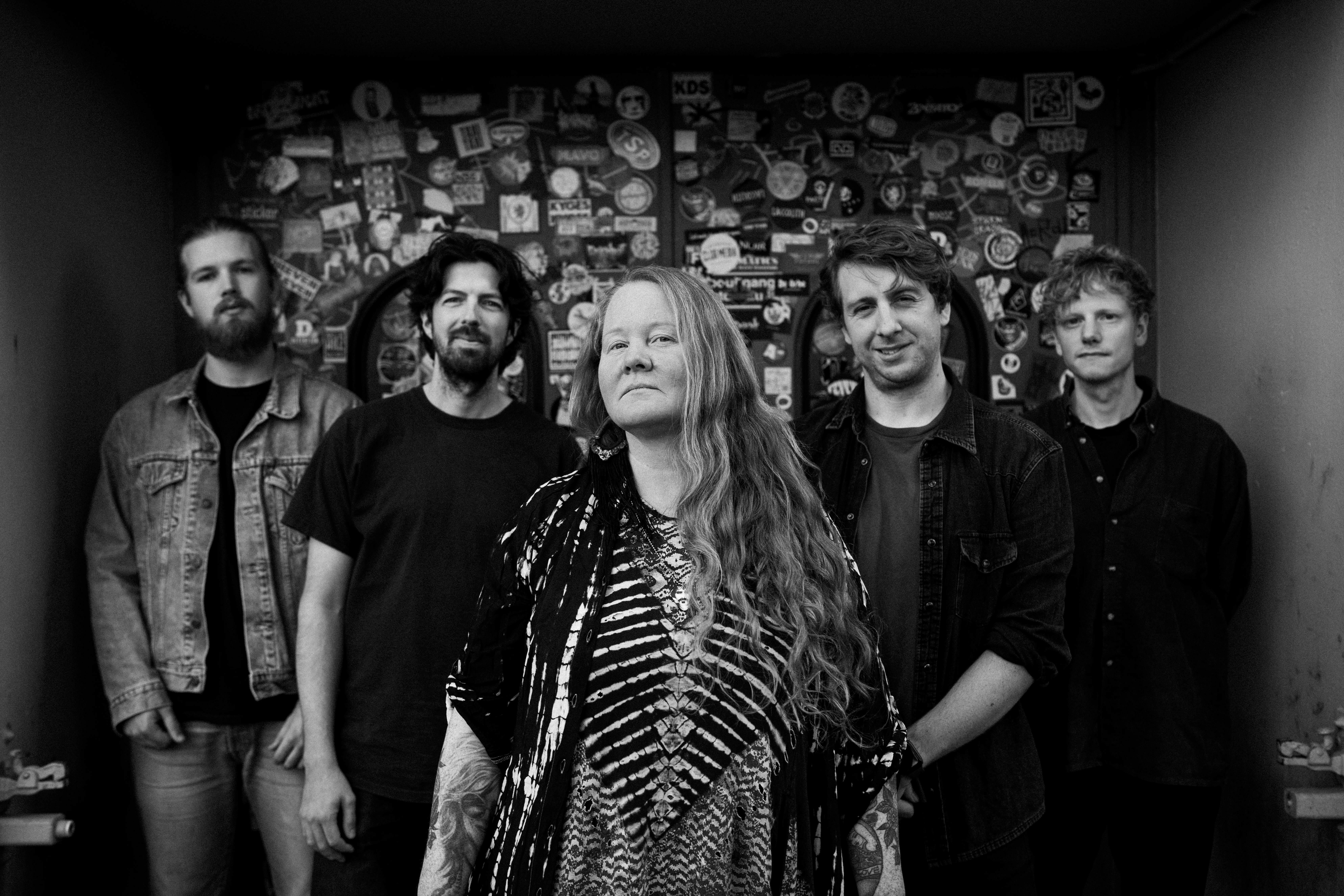 Schwarz-Weiß Foto der fünfköpfigen Band Layla Zoe vor einer Wand mit zahllosen Stickern und Aufklebern